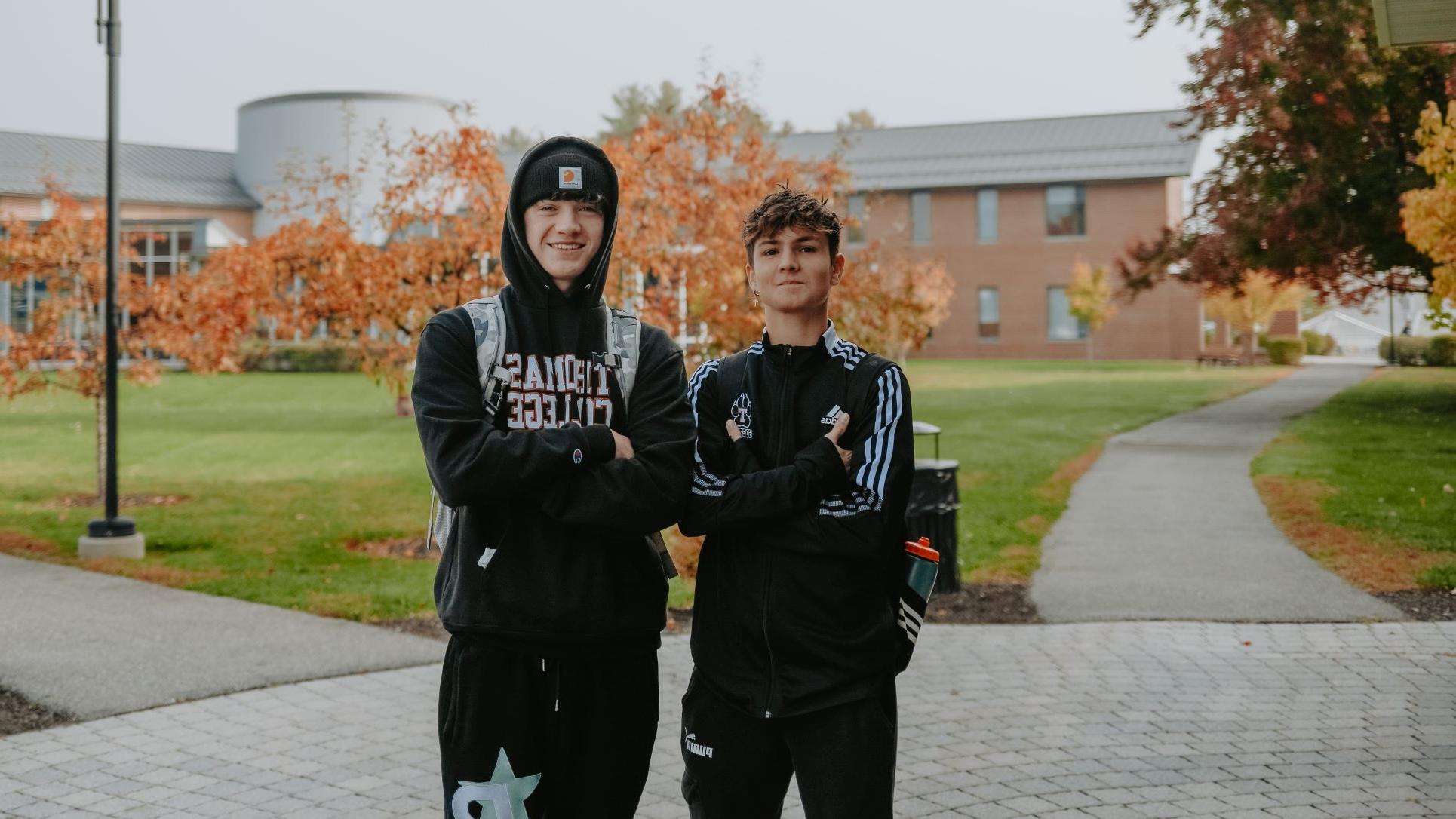 Two international students wearing Thomas College swag posed together with crossed arms on campus